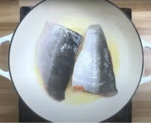Two salmon fillets skin-side down in a white pan on a stovetop, beginning to cook.